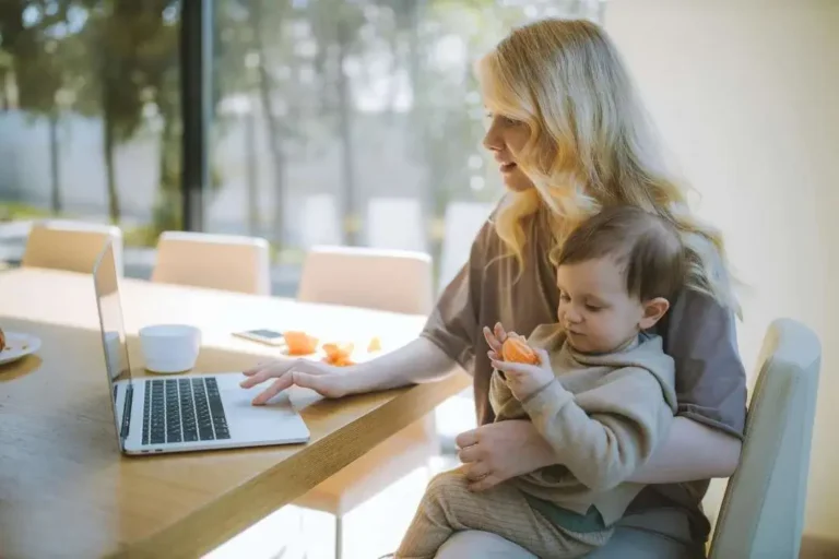 A woman researching on her laptop while holding a child who is enjoying a snack at the table.