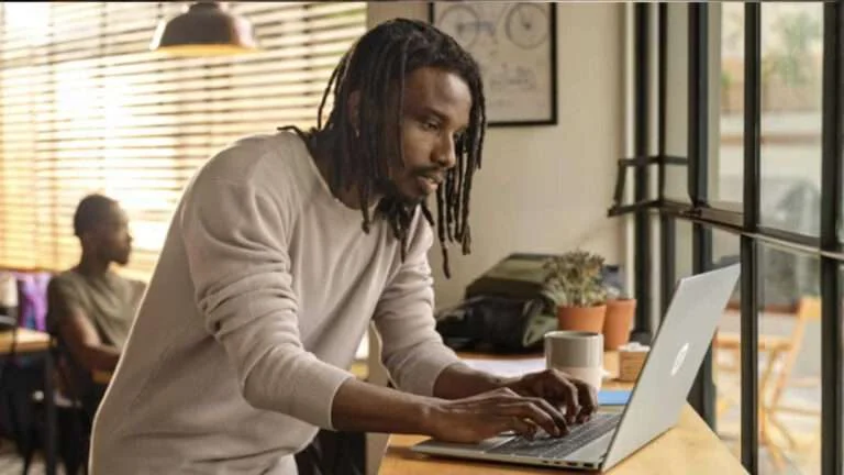 A person working on a laptop at a wooden counter in a well-lit room with large windows and a potted plant nearby.