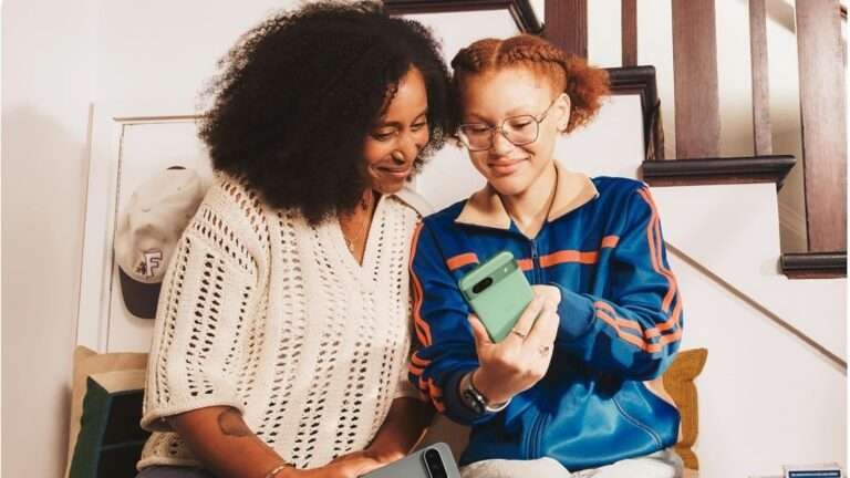 "Two women are sitting on a couch, one wearing a cream-colored knit sweater and the other in a blue and orange jacket, both holding and looking at their Pixel 9 phones.